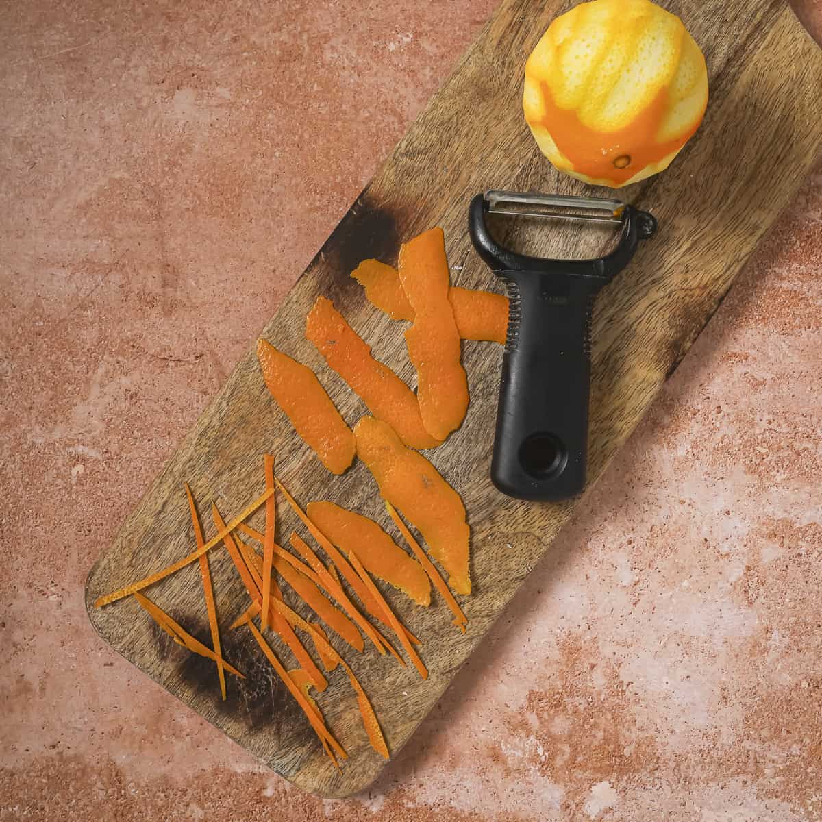 y-peeler and orange peel strips on a wooden cutting board.