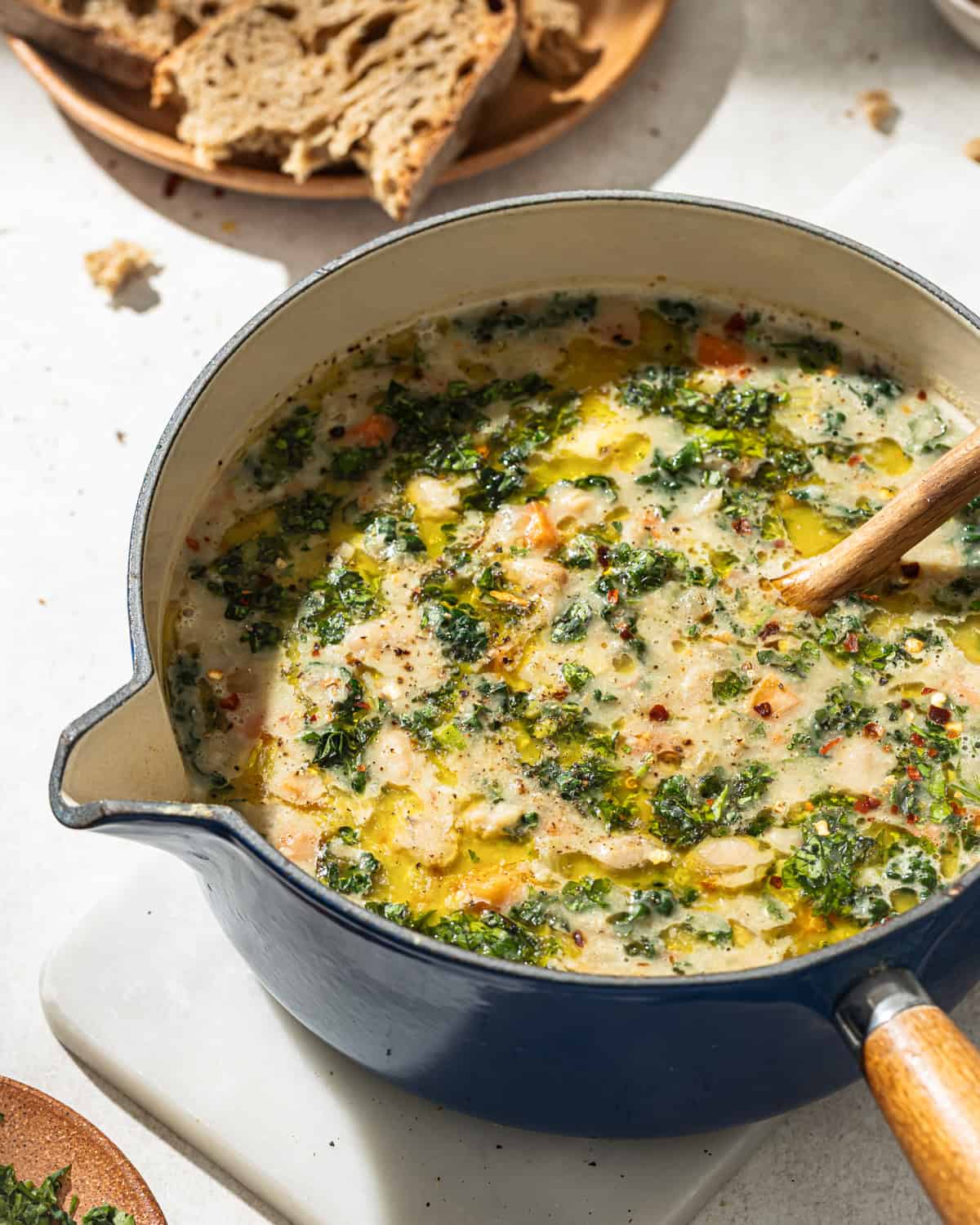 a blue pot of white bean soup with kale on a white surface with bread.