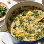 a blue pot of white bean soup with kale on a white surface.