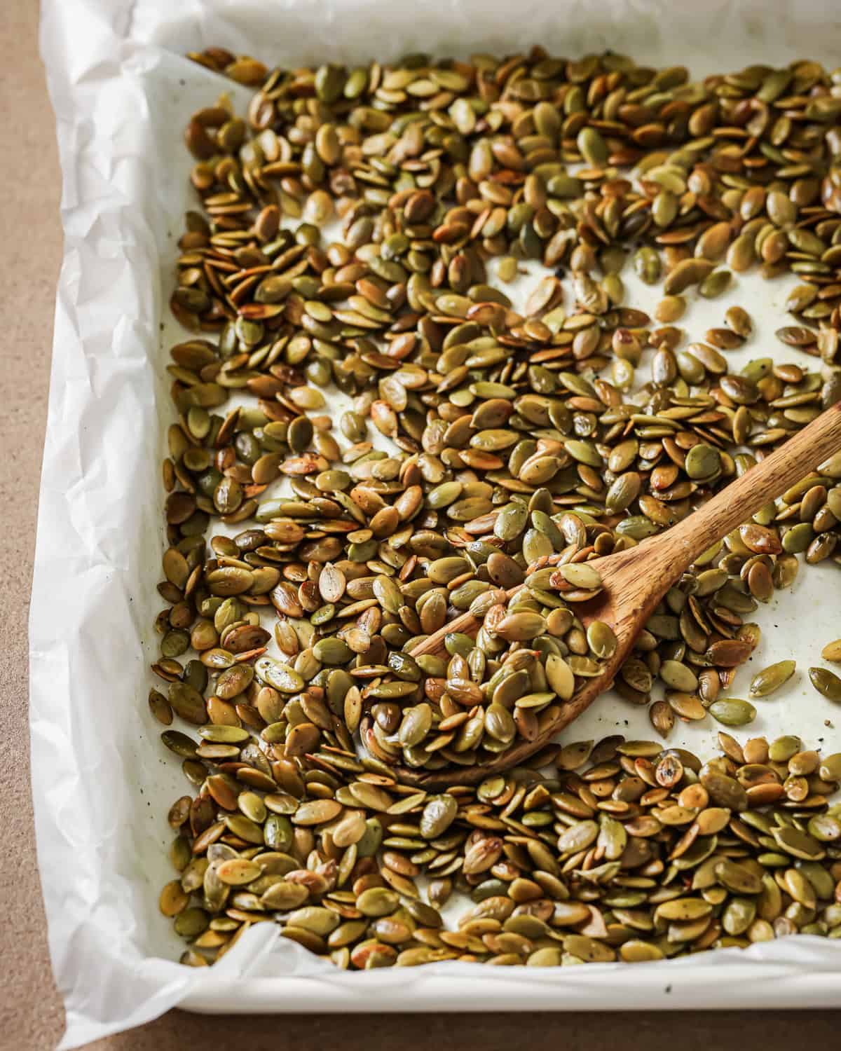 Roasted pepitas and a wooden spoon on a baking tray lined with parchment paper.