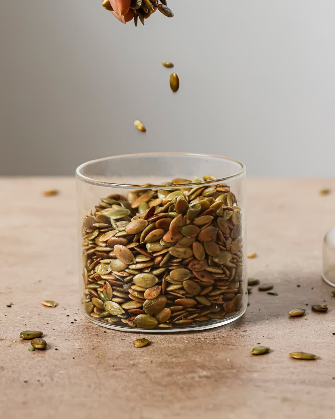 person sprinkling roasted pepitas into a glass jar on a table.