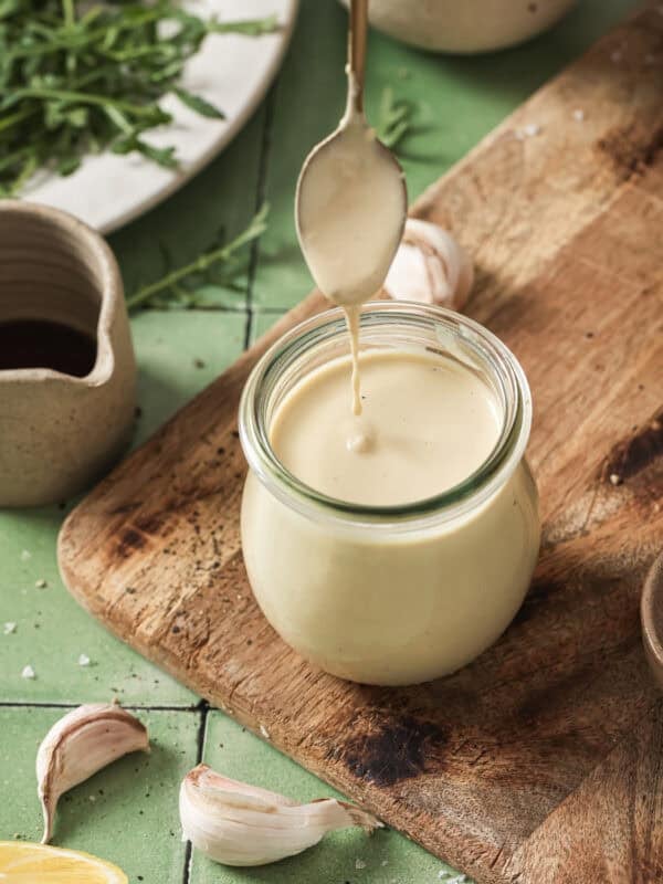 person removing spoon from jar of tahini dressing on a wooden cutting board.