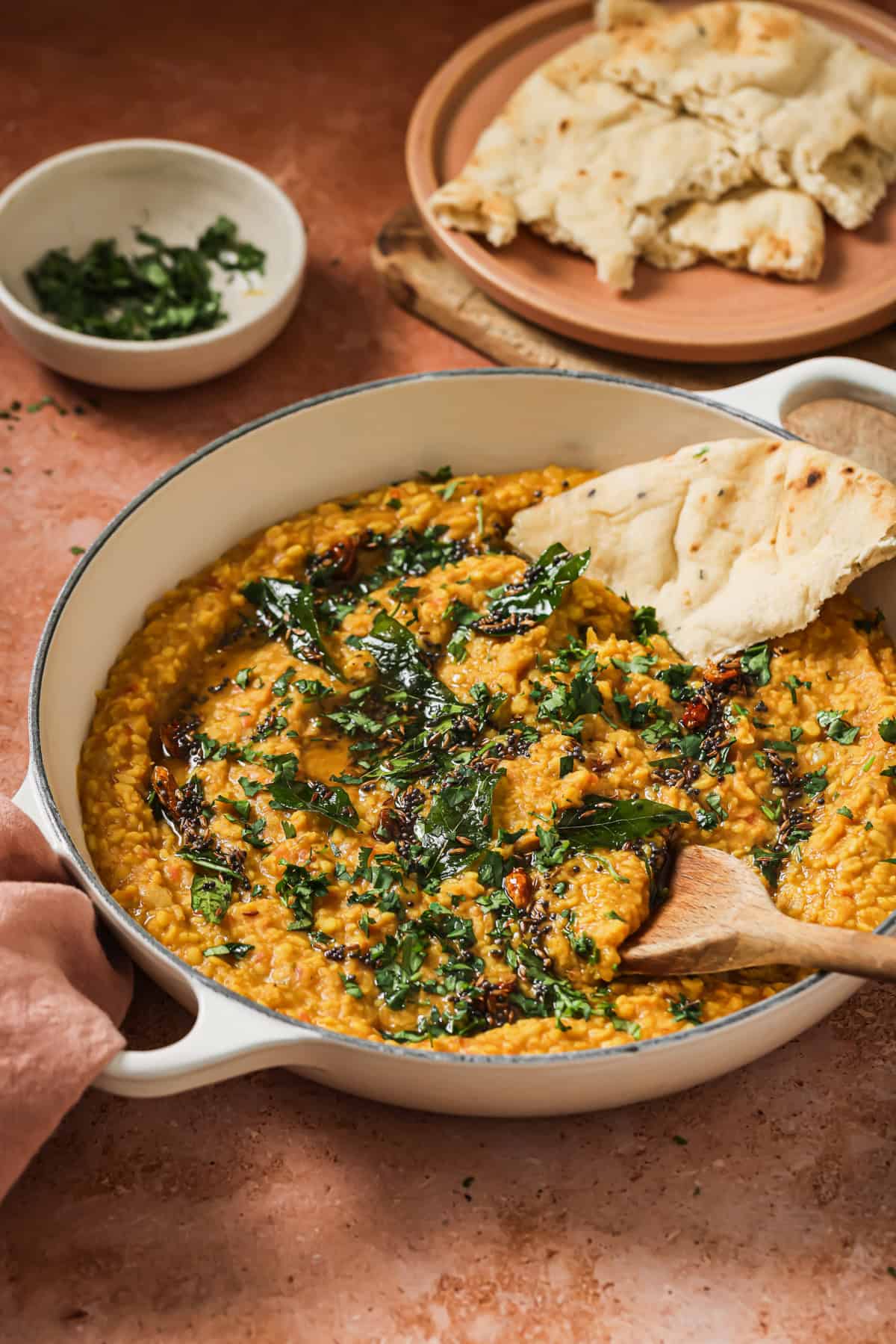 side view of dal tadka in deep skillet next to a plate of naan and bowl of cilantro.