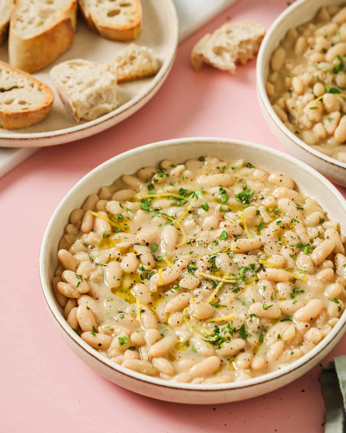a bowl of creamy white beans on a pink surface with bread in background.