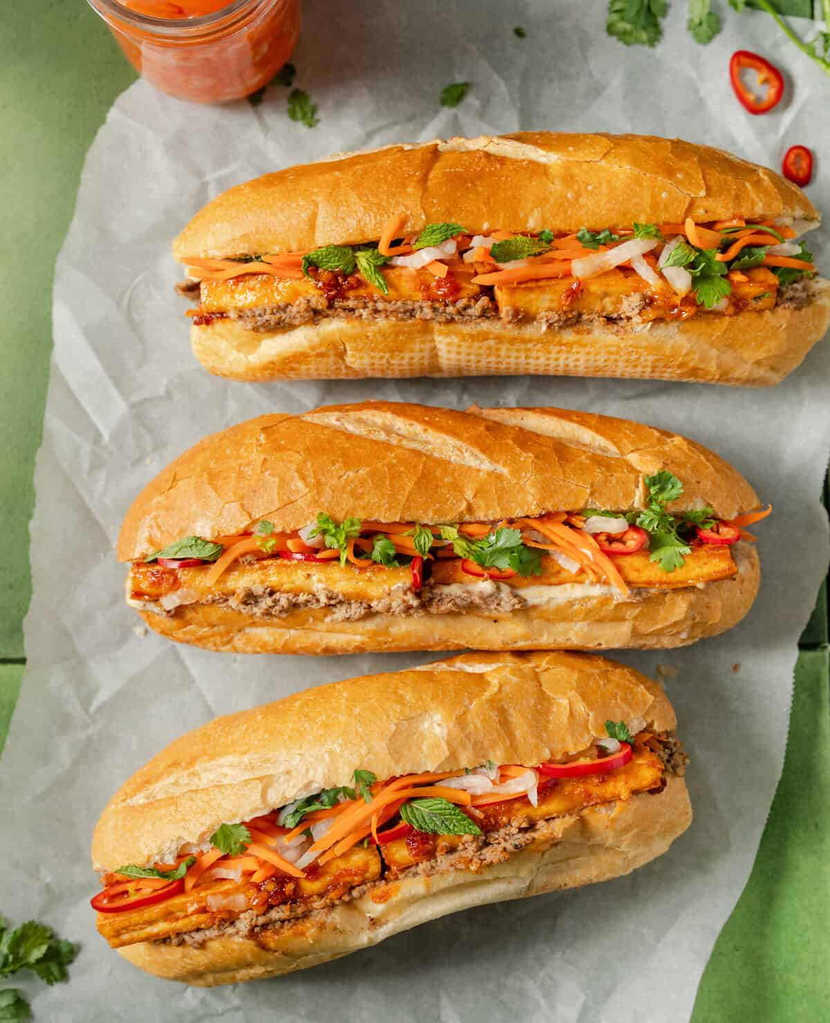 three tofu banh mi sandwiches arranged on parchment paper on a green tiled surface.