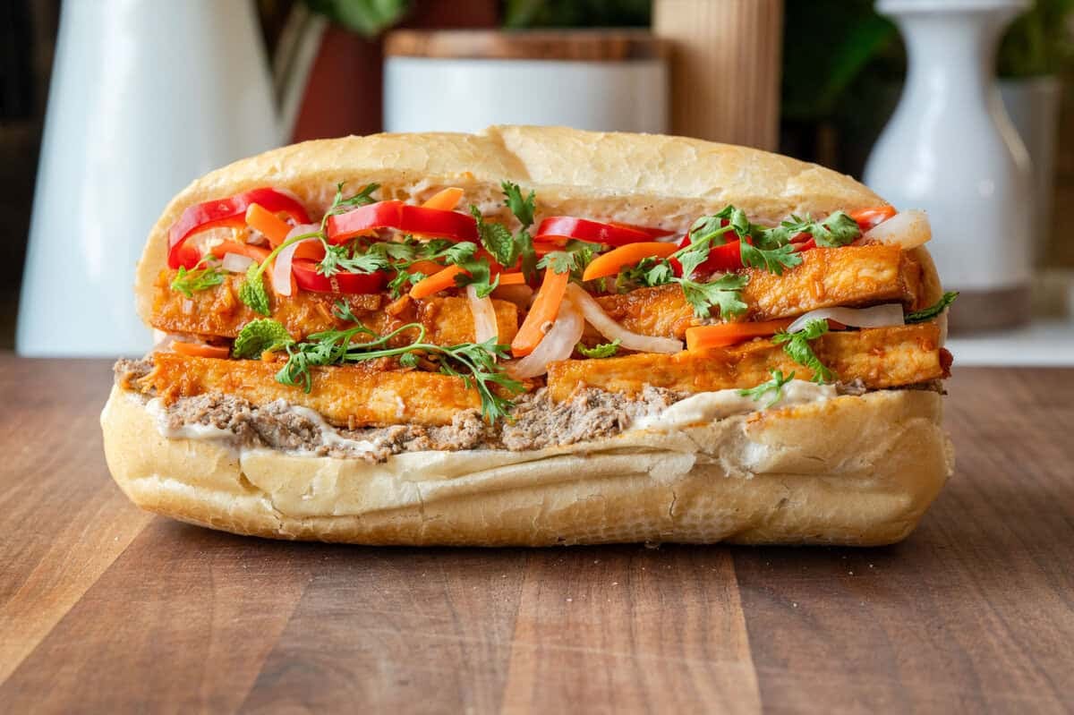 tofu banh mi loaded with toppings on a wooden cutting board.