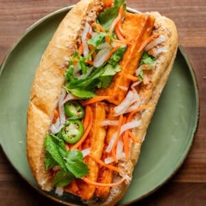 tofu banh mi loaded with toppings on a a green plate on a wooden cutting board.