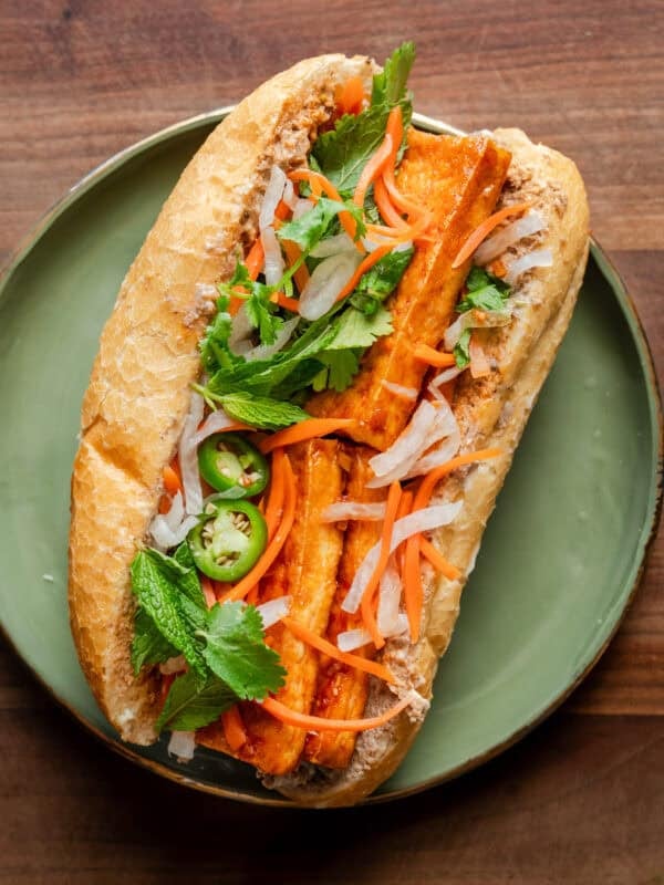 tofu banh mi loaded with toppings on a a green plate on a wooden cutting board.