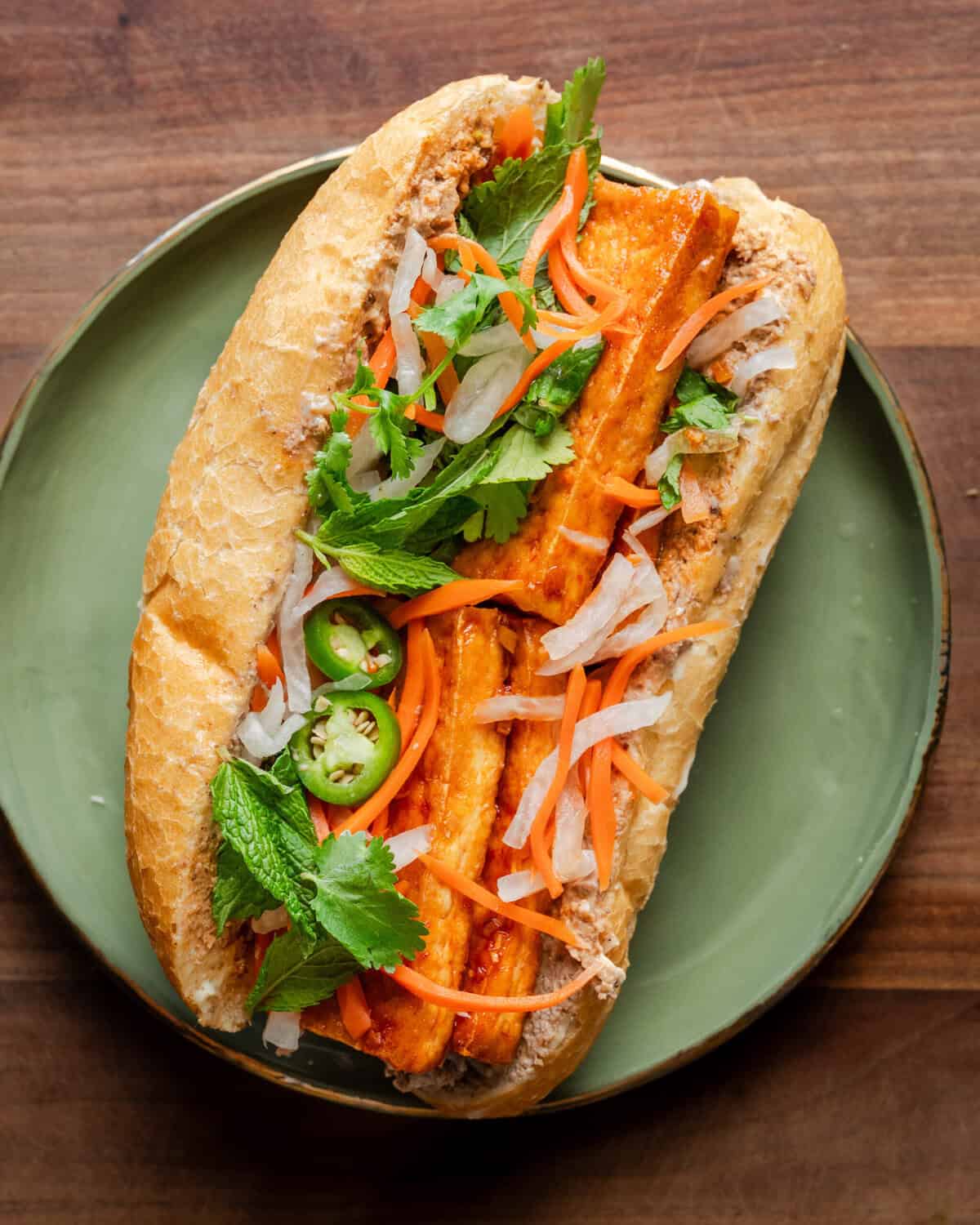 tofu banh mi loaded with toppings on a a green plate on a wooden cutting board.