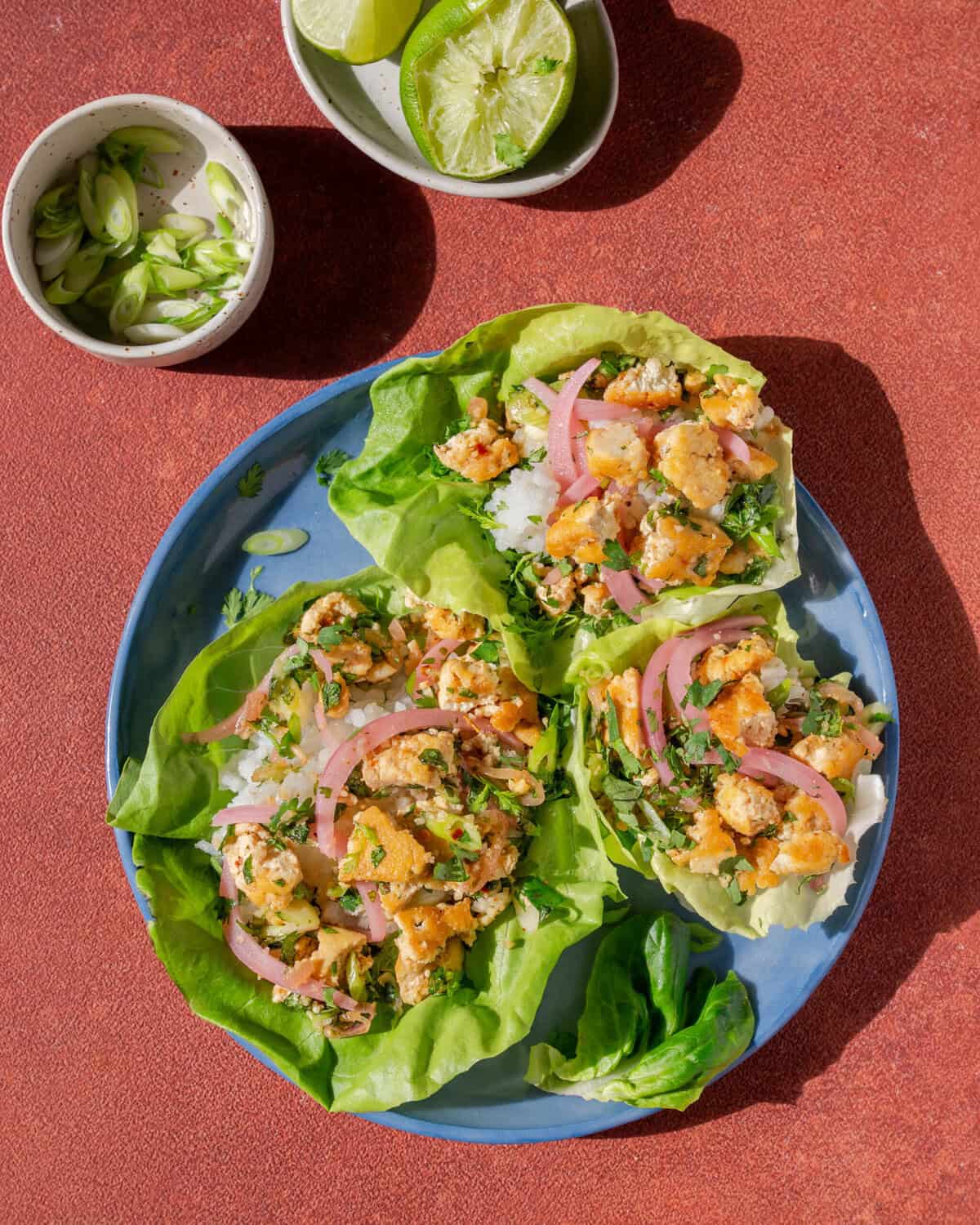three thai laab salad lettuce cups on a plate next to small bowls of lime and chilis.