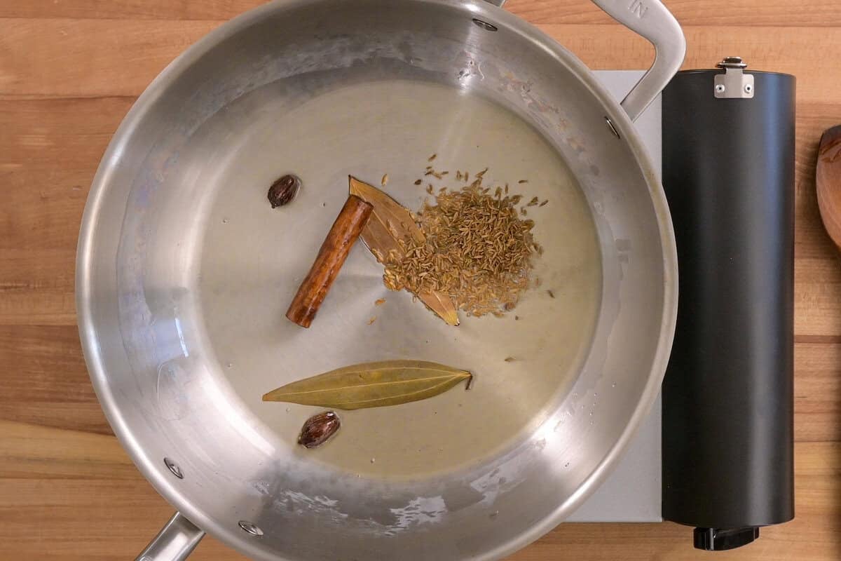 oil, cinnamon stick, bay leaves, cardamom and cumin cooking in a frying pan.