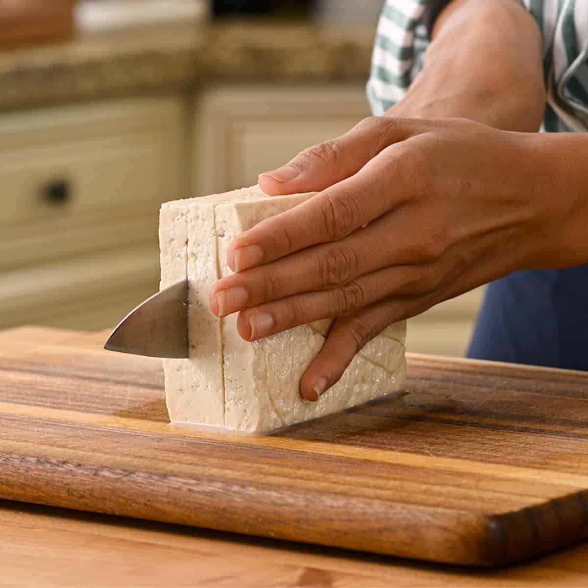 person slicing tofu into thin slabs on a wooden cutting board.