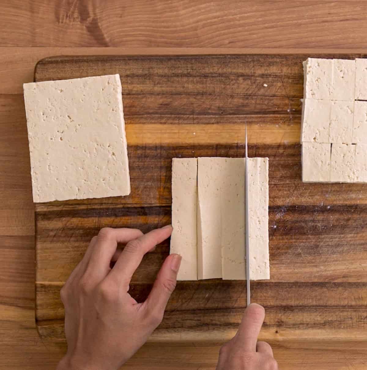 person slicing the tofu block vertically on a wooden cutting board.