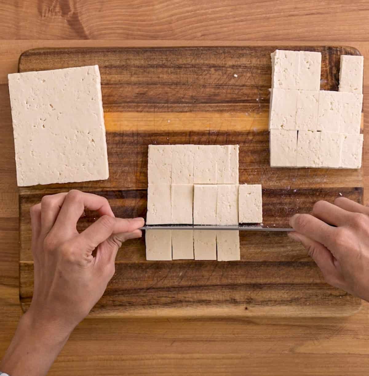 person slicing the tofu slabs crosswise on a wooden cutting board.